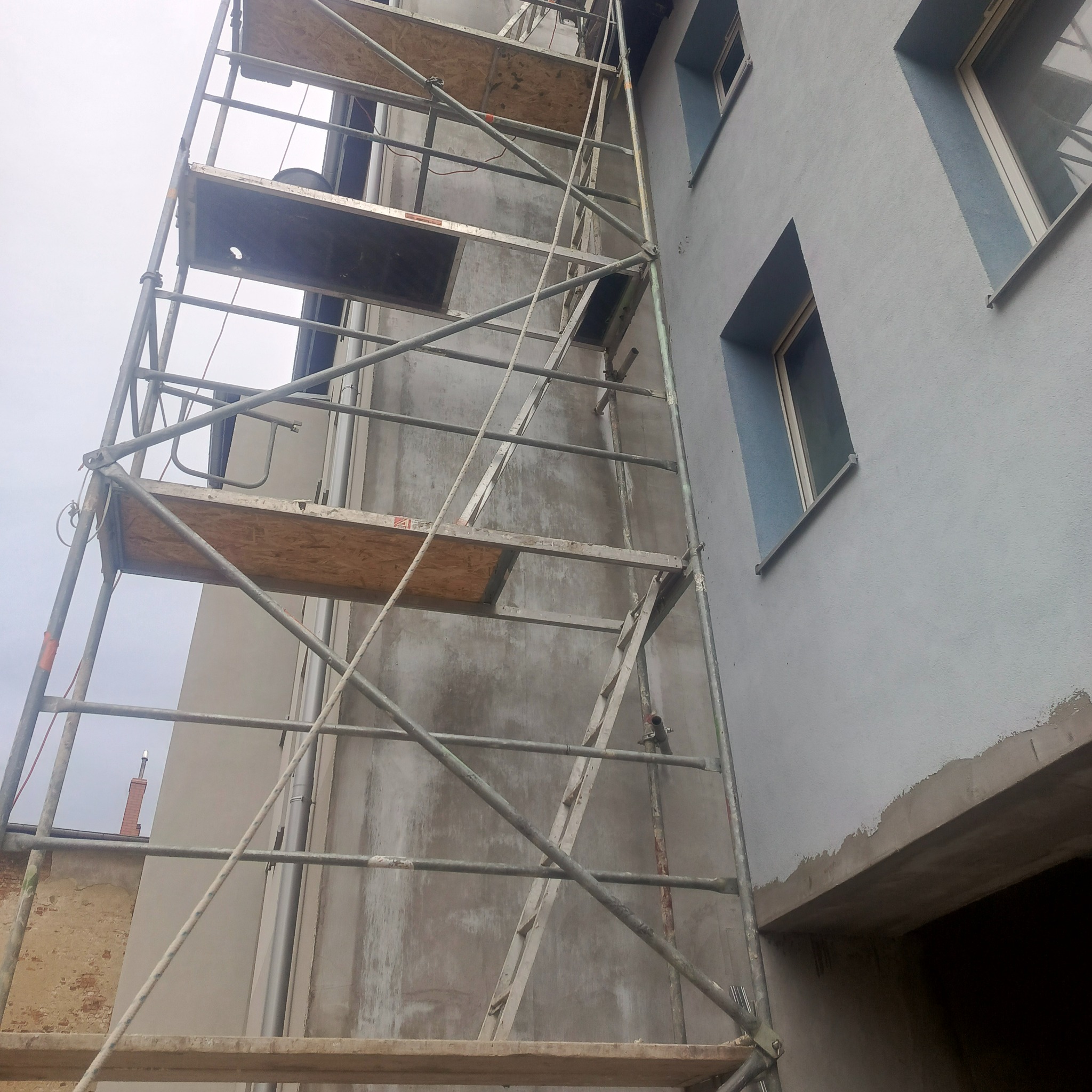Szare rusztowanie z drewnianymi podestami na tle błękitnej ściany budynku z oknami. Widoczny fragment elewacji w trakcie prac wykończeniowych.
