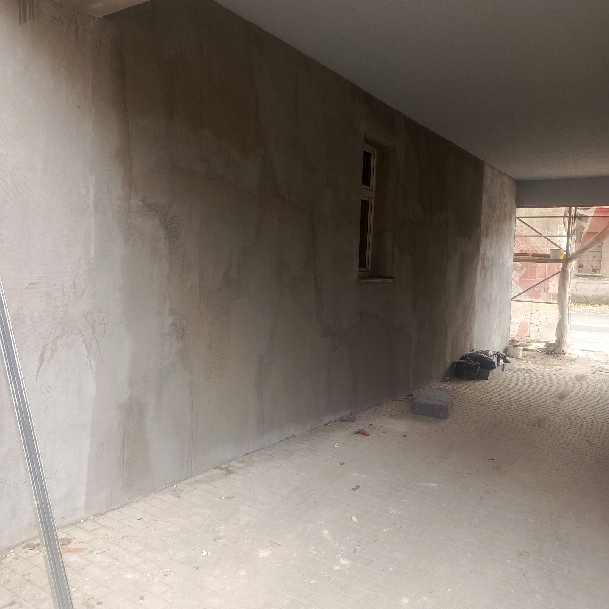 Ściana po nałożeniu szarego tynku, widoczne okno i fragment rusztowania. Prace wykończeniowe wewnątrz budynku, surowy stan.