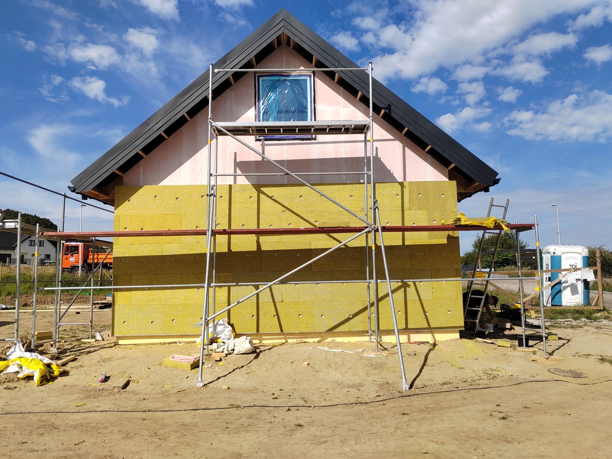 Ocieplanie budynku wełną mineralną, widoczne rusztowanie i okno zabezpieczone folią. Prace w toku, budynek z czarnym dachem na tle błękitnego nieba.