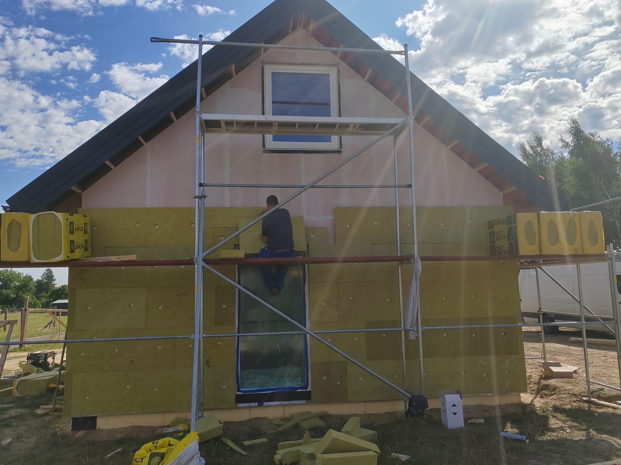 Ocieplanie elewacji domu jednorodzinnego z użyciem rusztowania. Widoczne płyty izolacyjne z wełny mineralnej, częściowo zamontowane. Prace w toku, słoneczny dzień.