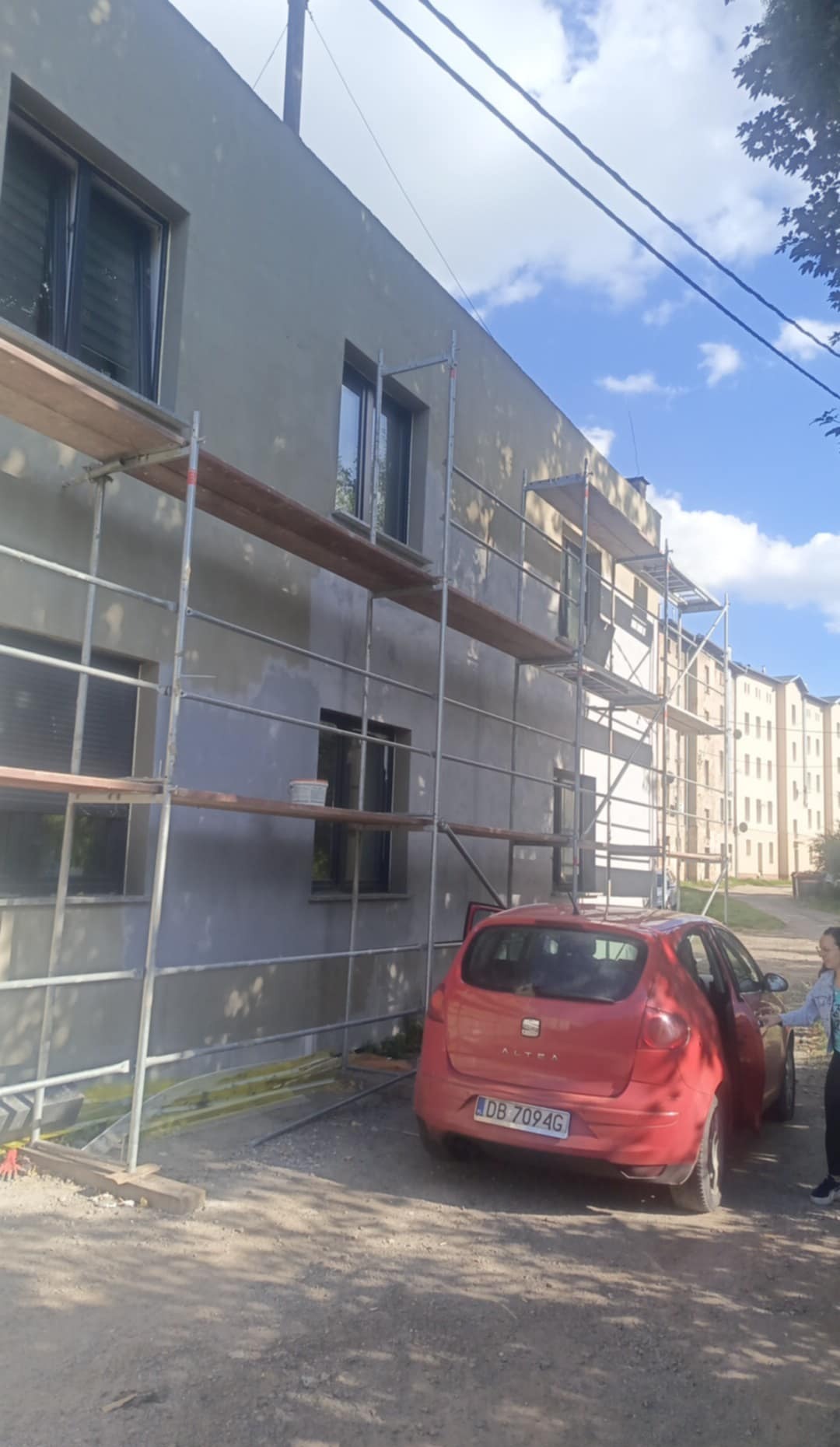 Budynek w trakcie prac elewacyjnych, widoczne rusztowania, częściowo otynkowana ściana, czerwony samochód osobowy i fragment sylwetki kobiety.
