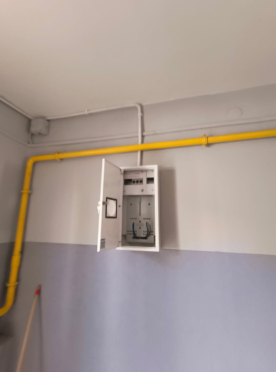 Otwarta skrzynka z instalacją elektryczną na dwukolorowej ścianie, z żółtą rurą gazową biegnącą wzdłuż sufitu. Widoczne przewody i elementy instalacji.