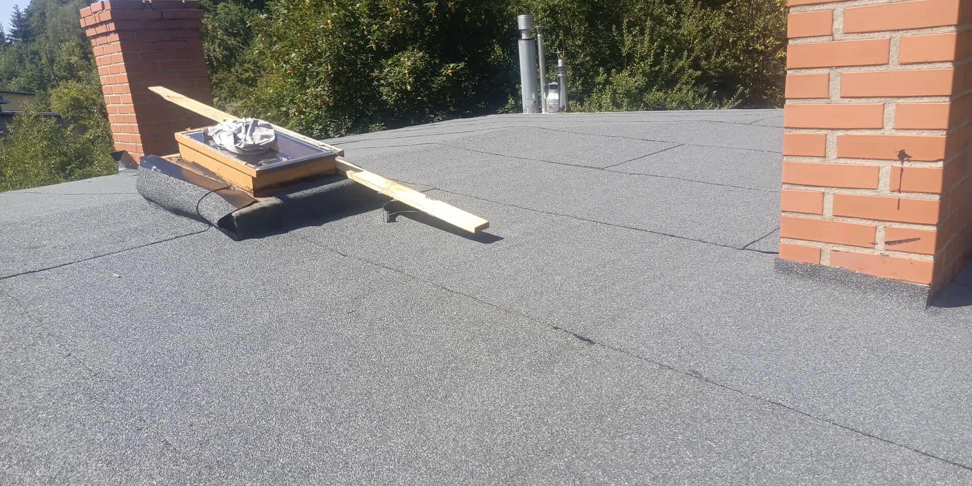 Szary dach pokryty papą z kominem z cegły i oknem dachowym w trakcie montażu. Widoczne narzędzia i elementy konstrukcyjne. Słoneczny dzień.