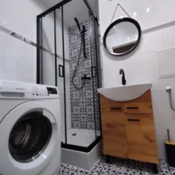 Wykończona łazienka w bieli i czerni z kabiną prysznicową z czarną ramą, umywalką na drewnianej szafce, pralką i dekoracyjnymi płytkami podłogowymi.