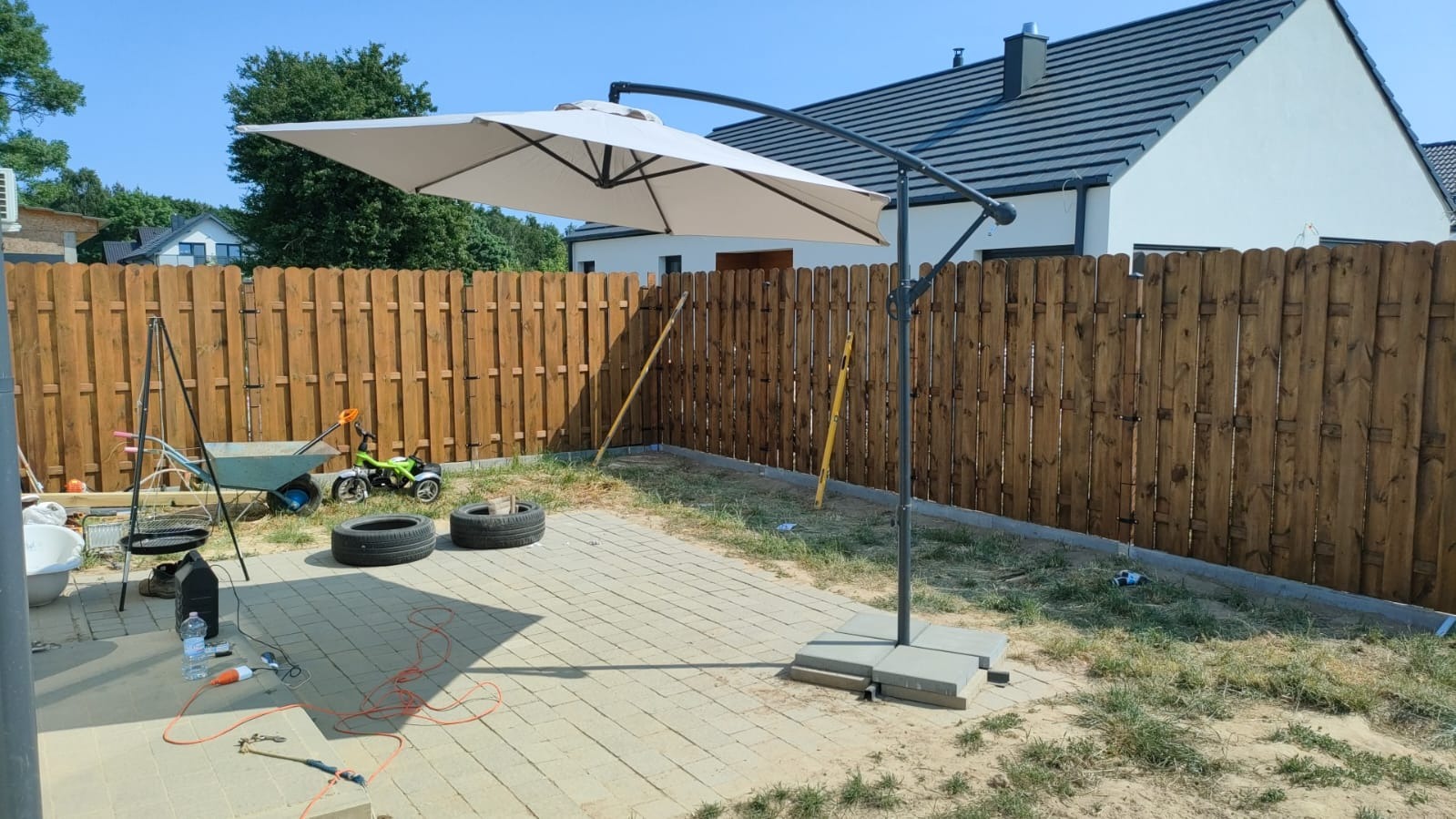 Nowy, drewniany płot lamelowy z brązowym impregnatem, widok zza rozłożonego parasola ogrodowego na betonowej kostce, w tle biały dom z ciemną dachówką.