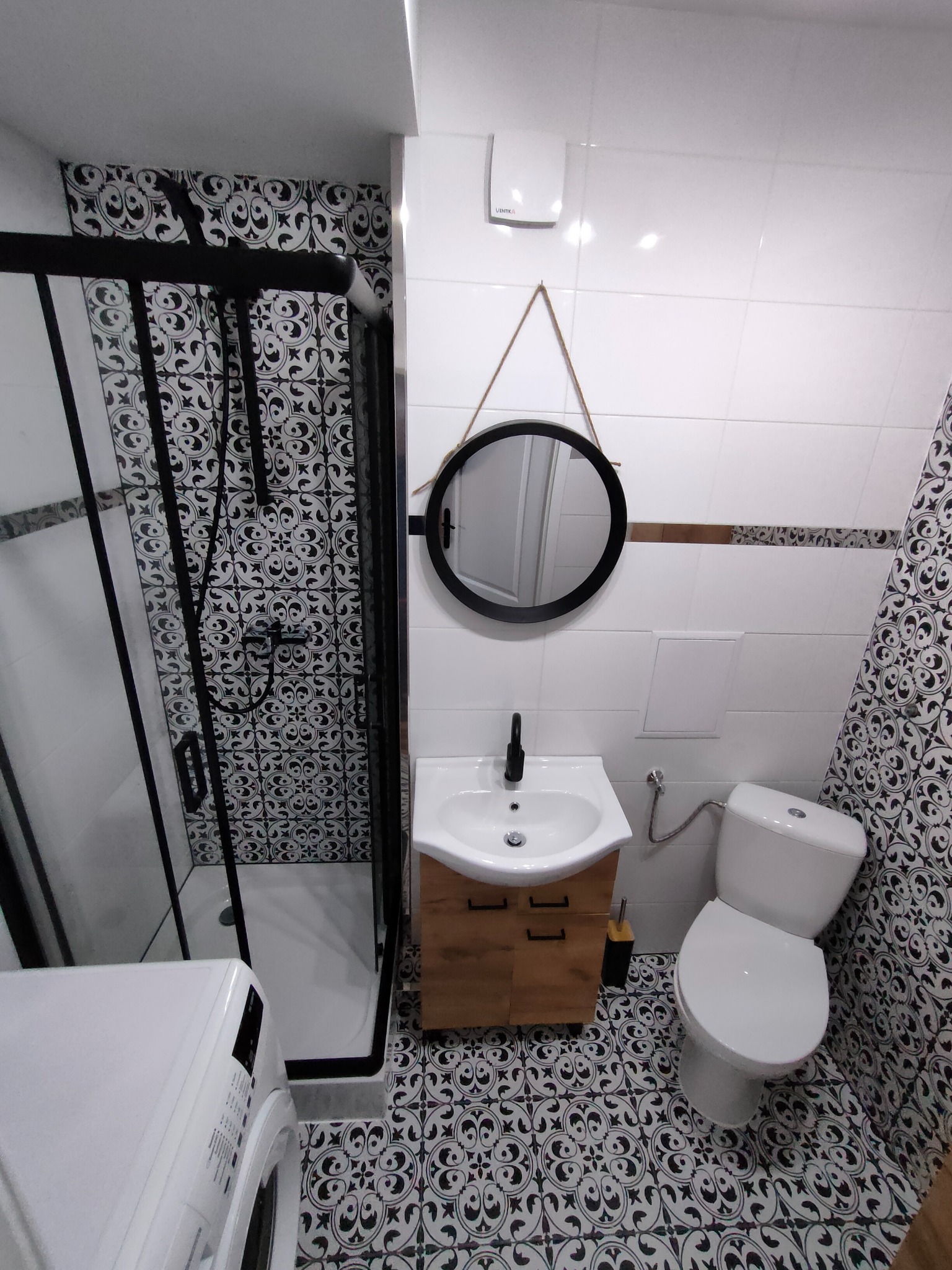 Wykończona łazienka z kabiną prysznicową typu walk-in z czarnymi ramami, umywalką z szafką z drewna, okrągłym lustrem na sznurku, toaletą i pralką, z dekoracyjnymi czarno-białymi płytkami...