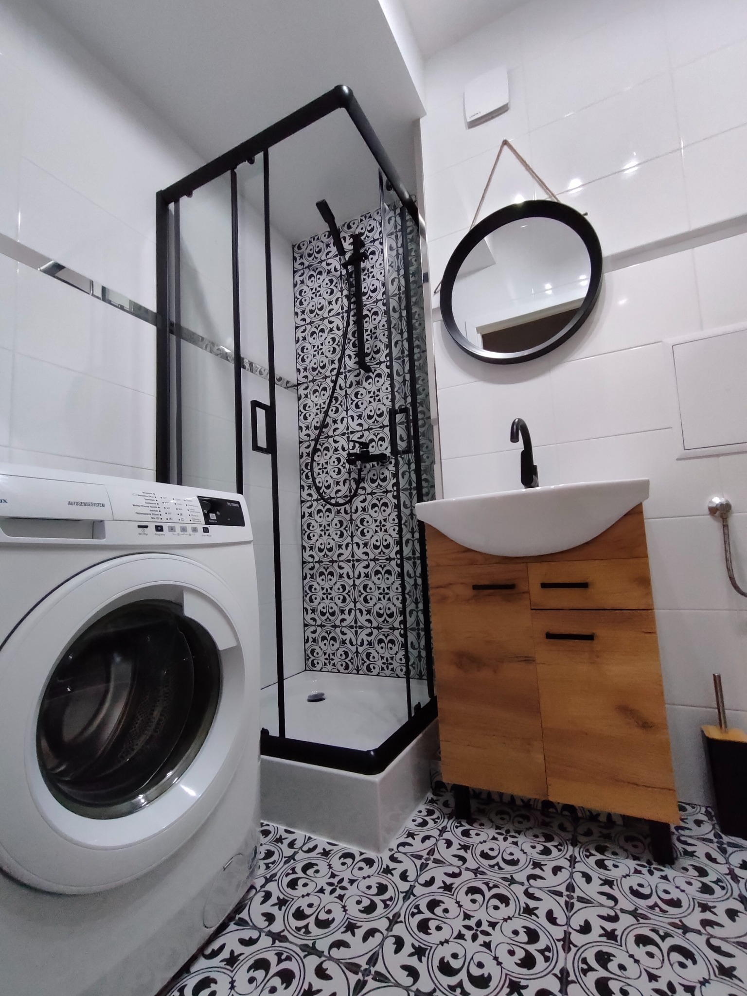 Wykończona łazienka w bieli i czerni z kabiną prysznicową z czarną ramą, umywalką na drewnianej szafce, pralką i dekoracyjnymi płytkami podłogowymi.
