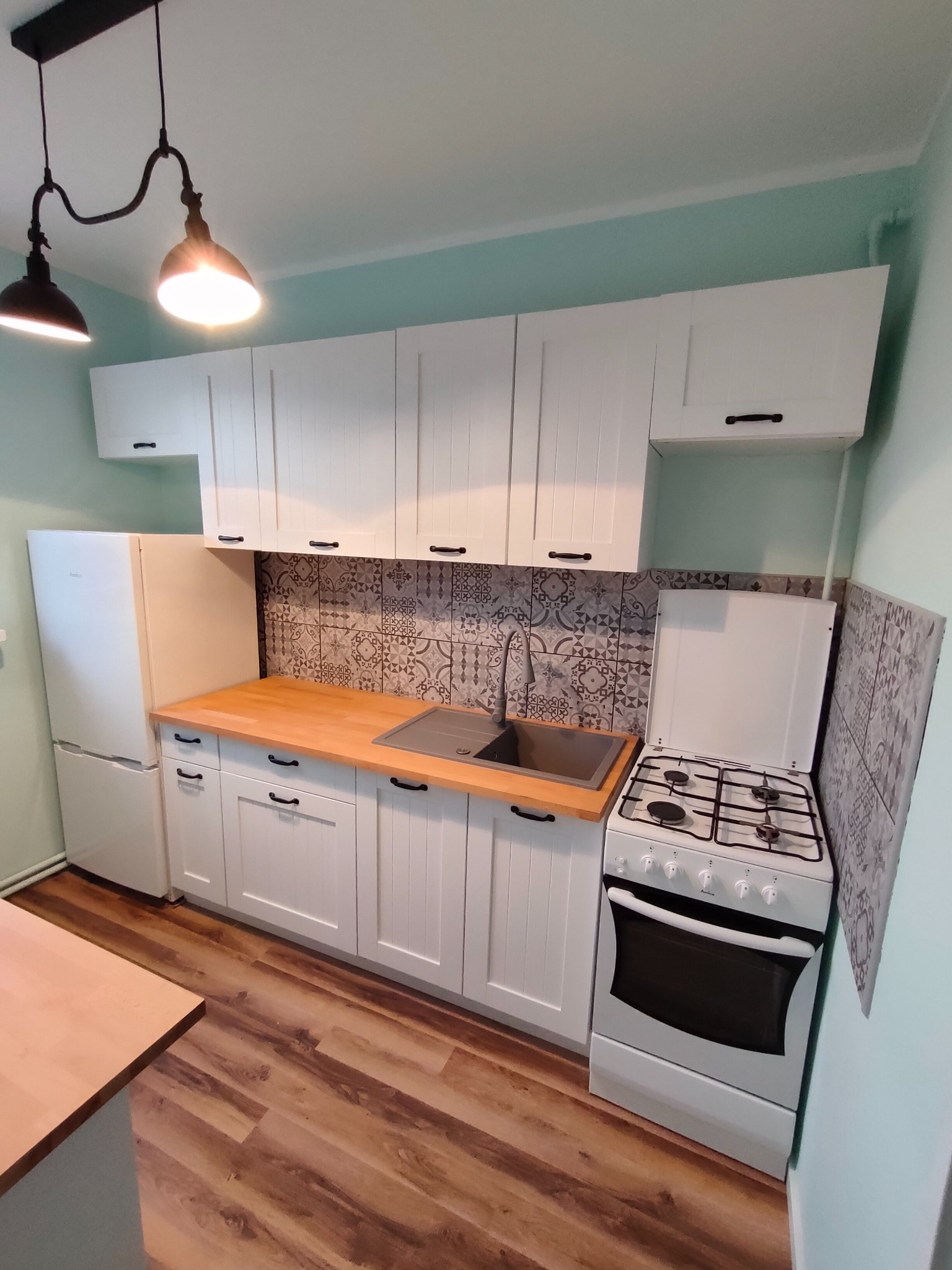 Jasna kuchnia z białymi szafkami, drewnianym blatem i wzorzystą mozaiką nad zlewem, oświetlona lampą w stylu industrialnym.