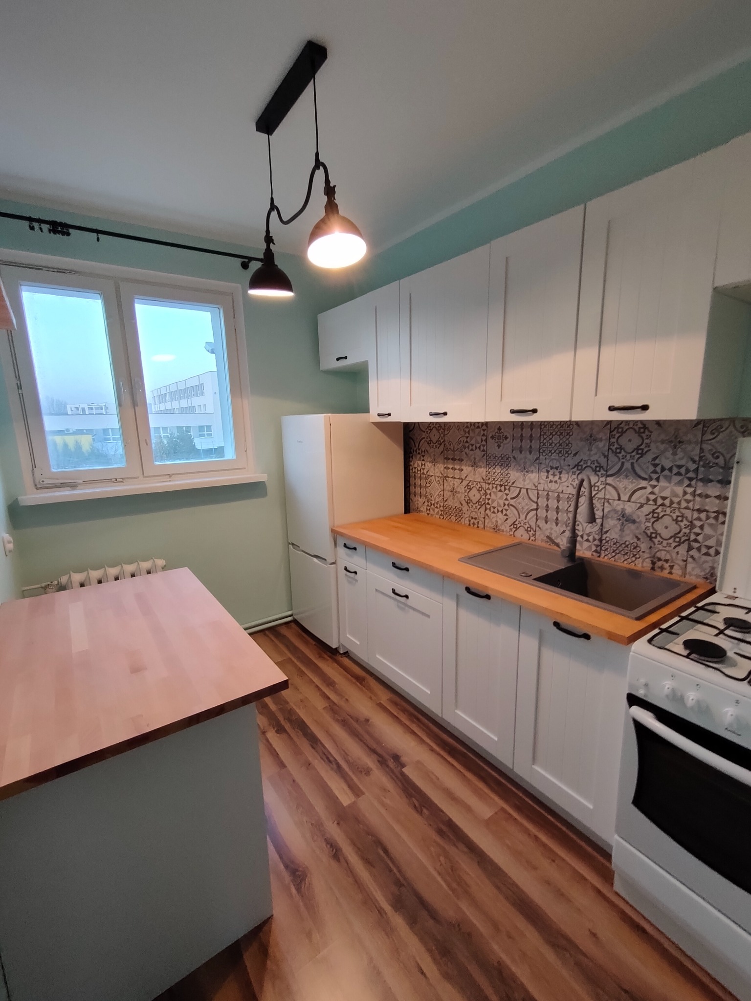 Jasna kuchnia z białymi szafkami, drewnianym blatem i wzorzystą mozaiką nad zlewem. Na suficie designerska lampa z dwoma kloszami. Widok z okna na budynek mieszkalny.