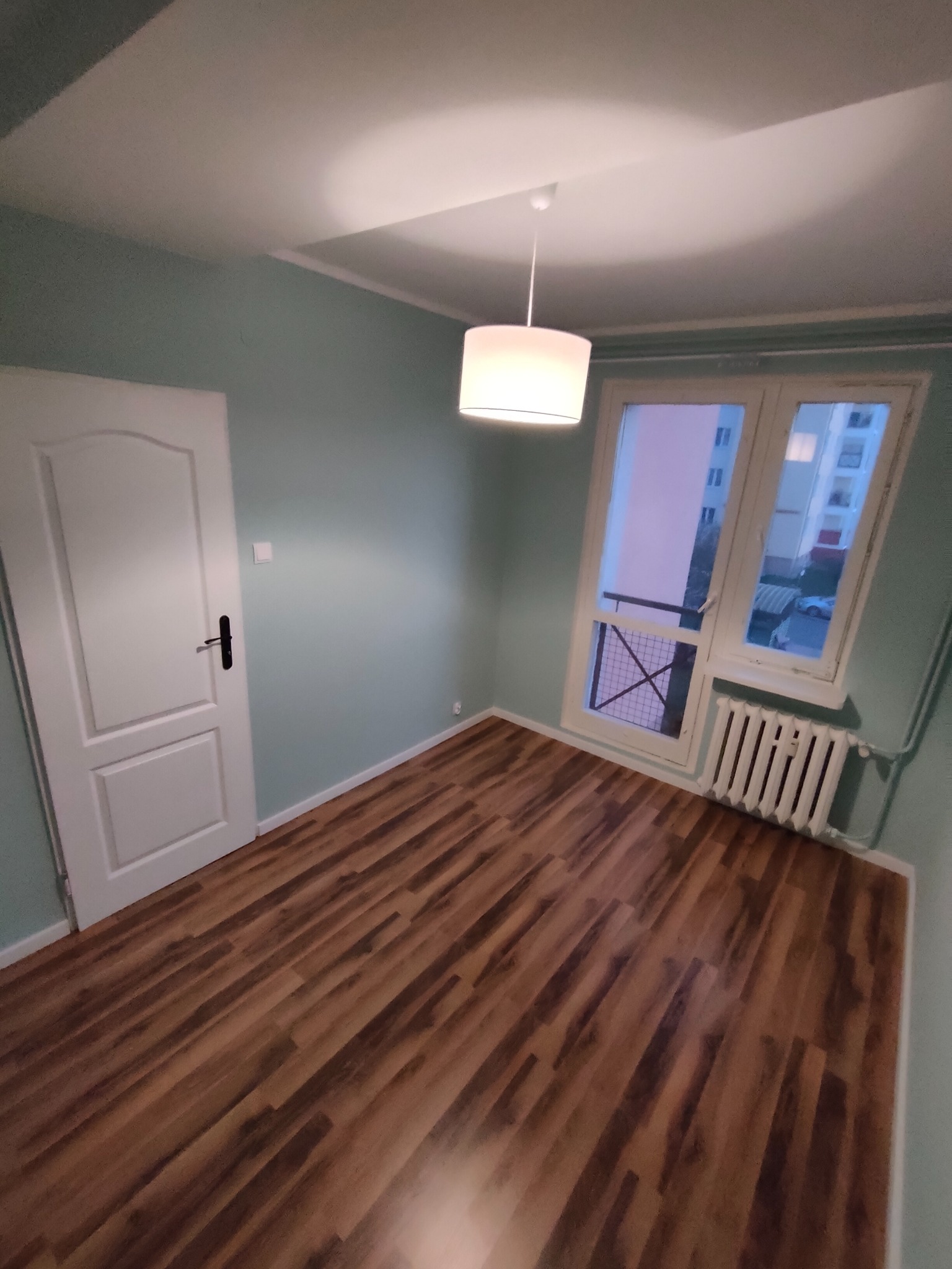 Pomalowane na jasny, pastelowy odcień zieleni ściany w pustym pokoju z panelami podłogowymi, białe drzwi z czarną klamką, okno z grzejnikiem i wisząca lampa z białym abażurem.