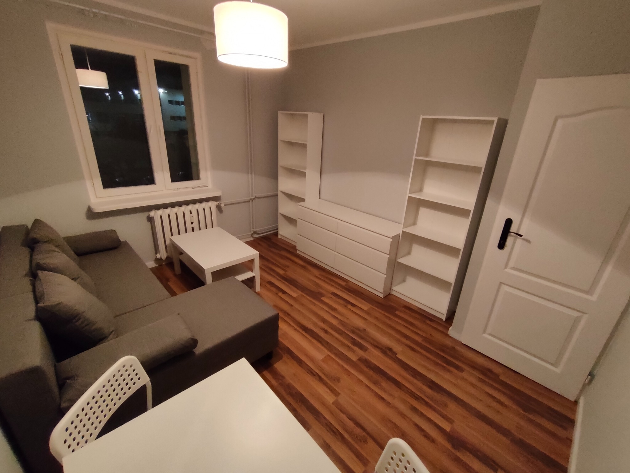 Nowoczesny salon z szarą sofą narożną, białymi meblami modułowymi, drewnianą podłogą i białymi krzesłami przy stole, oświetlony lampą sufitową.