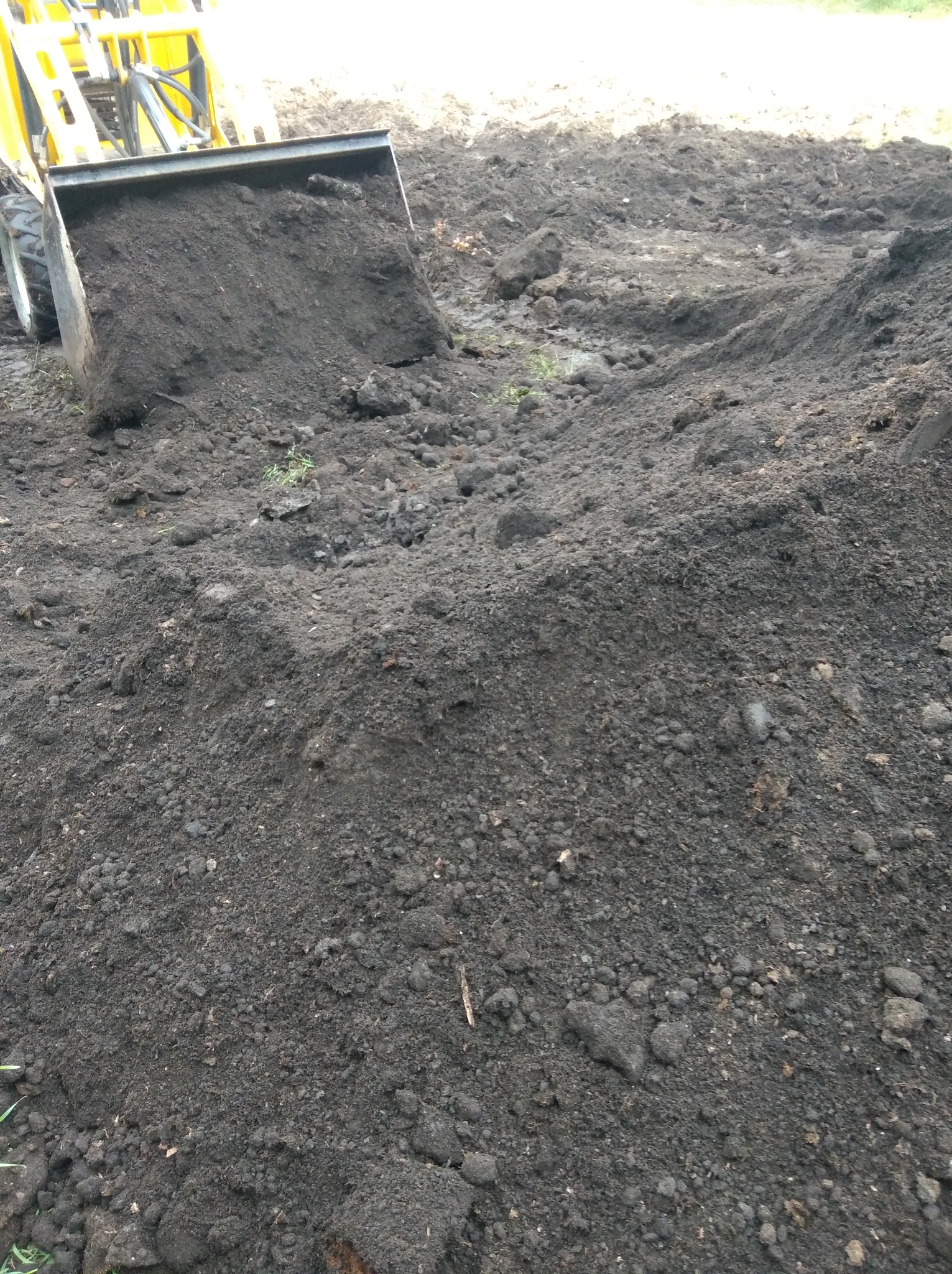 Żółta miniładowarka z łyżką wypełnioną ciemną ziemią na nierównym terenie. Widoczne ślady pracy w ziemi, częściowo zielona trawa. Sprzęt budowlany w trakcie wyrównywania terenu.