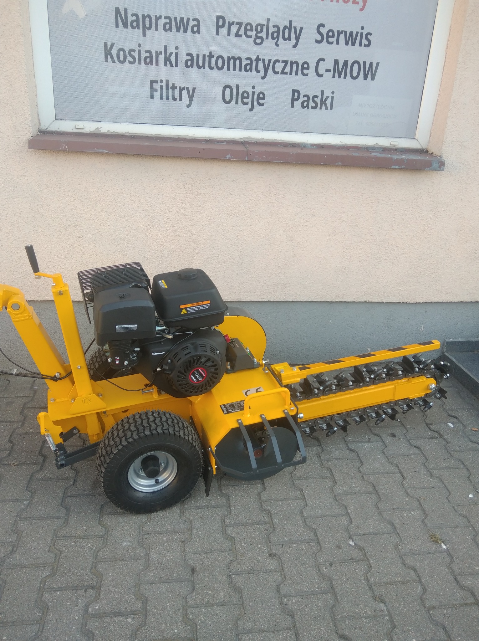 Żółta glebogryzarka łańcuchowa na kostce brukowej, z widocznym silnikiem i ostrzami. W tle ściana budynku z napisem 'Kosiarki automatyczne C-MOW'.