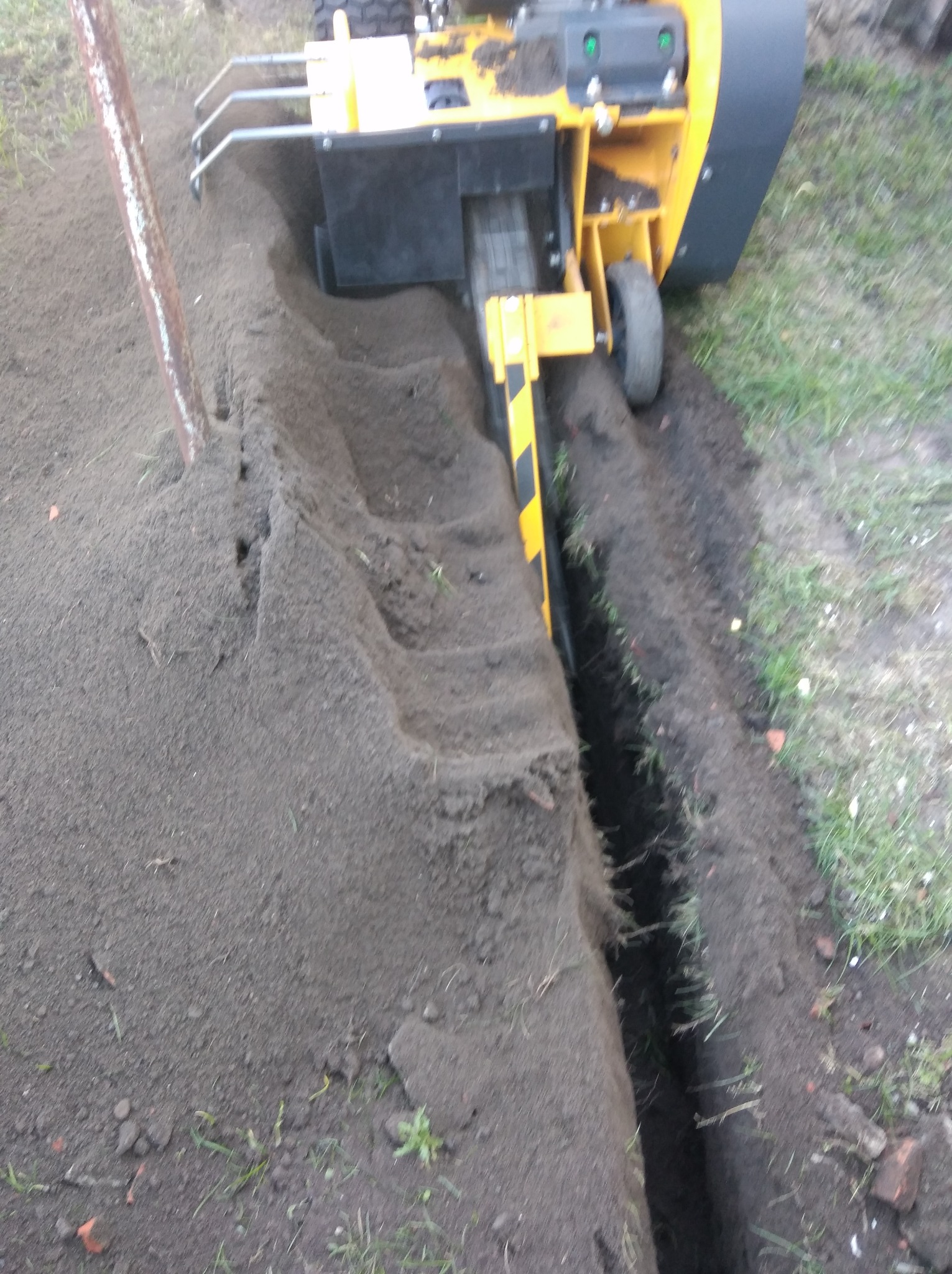 Żółta maszyna wykopuje wąski rów w ziemi, przygotowując teren pod instalację systemu nawadniania. Widoczny świeżo wykopany rów i pryzma ziemi obok.