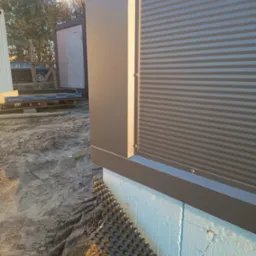 Fragment budynku z szarą blachą trapezową, widoczne mocowania, ocieplenie ze styropianu i folia kubełkowa przy gruncie, na zewnątrz teren budowy.