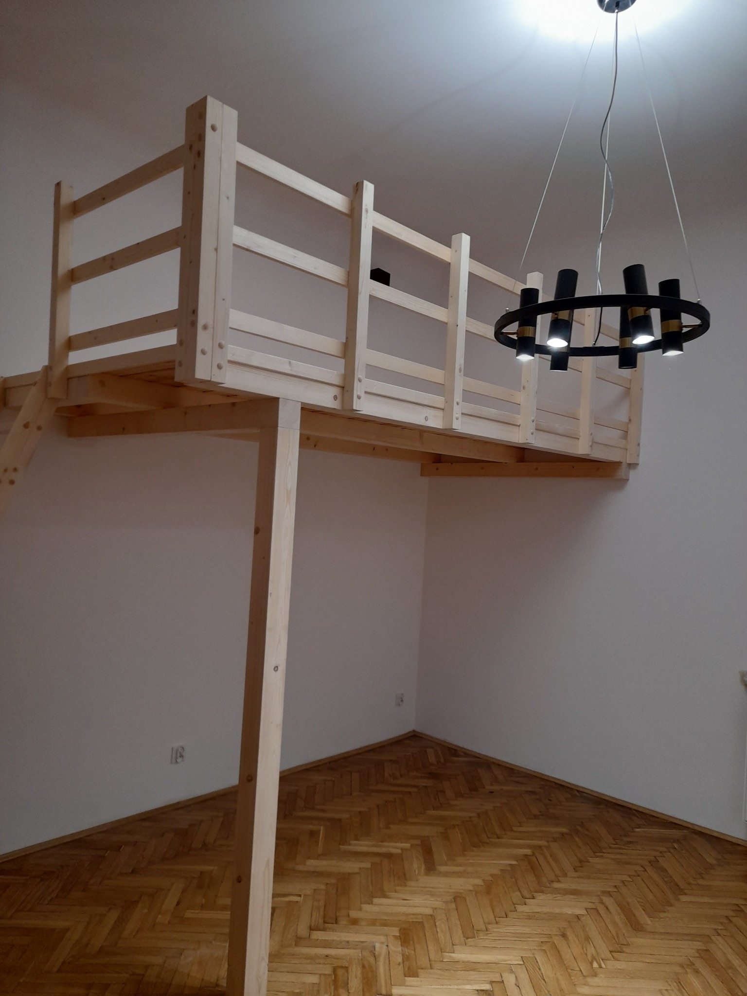 Drewniane łóżko na antresoli z drabinką w pokoju z parkietem i nowoczesnym okrągłym żyrandolem.