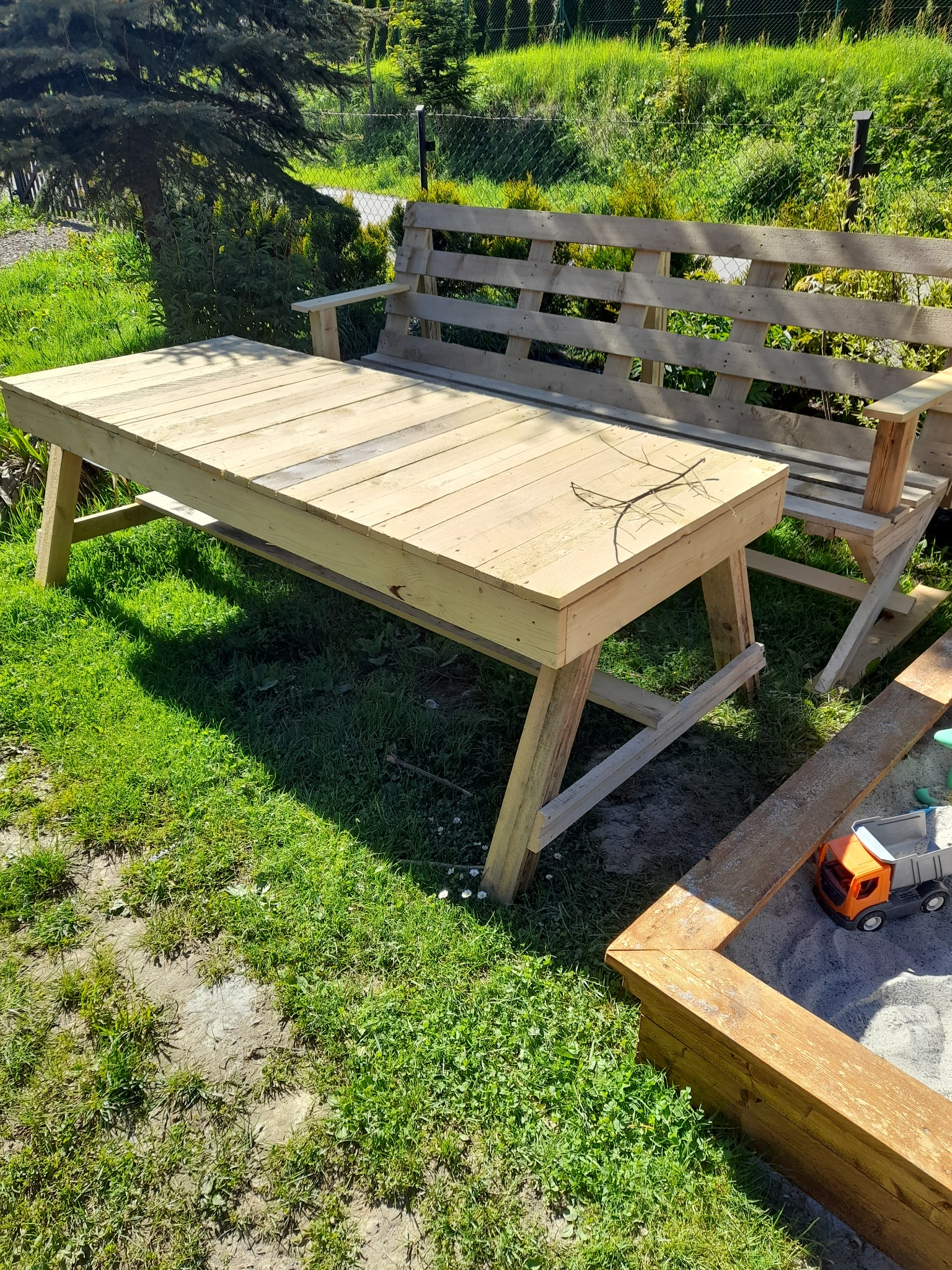 Drewniany stół i ławka ogrodowa wykonane z palet, ustawione na trawniku obok piaskownicy z zabawką, w tle zielona roślinność.