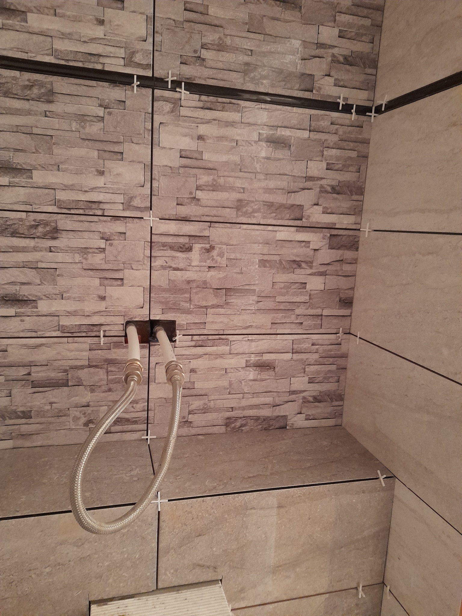 Ściana wyłożona płytkami imitującymi kamień, z widocznymi rurami i krzyżykami dystansowymi, w trakcie montażu.