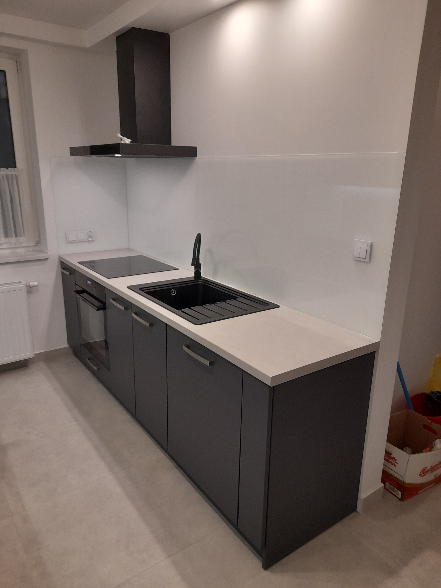 Nowoczesna, szara zabudowa kuchenna z czarnym zlewem i płytą indukcyjną, biały panel szklany nad blatem.