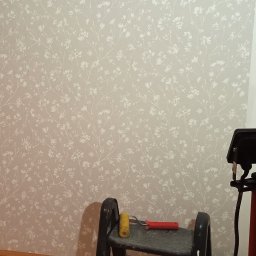Usługi remontowe malowanie, panele, płytki. - Ściana po częściowym pomalowaniu z wzorem kwiatowym, obok drabina z wałkiem i lampa robocza na statywie, widoczny fragment gniazdka elektrycznego przy podłodze.
