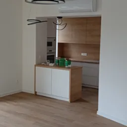 Jasne wnętrze mieszkania z podłogą z paneli w odcieniach drewna, widok na balkon i otwartą kuchnię z drewnianymi szafkami, nowoczesne oświetlenie.