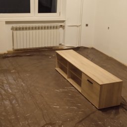 Usługi remontowe malowanie, panele, płytki. - Pokój w trakcie przygotowań do prac wykończeniowych, podłoga zabezpieczona folią ochronną, widoczny grzejnik i częściowo umeblowany szafką RTV z jasnego drewna.