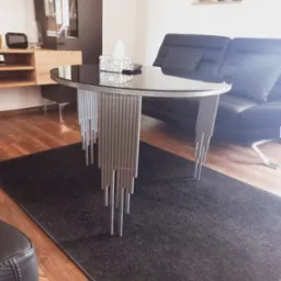 Okrągły stolik kawowy z czarnym szklanym blatem i srebrnymi, rurkowymi nogami stoi na ciemnoszarym dywanie w salonie z drewnianą podłogą i szarą sofą.