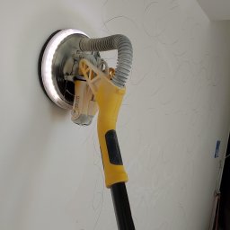 Usługi remontowe malowanie, panele, płytki. - Szlifierka do gładzi gipsowych Stanley Fatmax z oświetleniem LED na ścianie z widocznymi śladami szlifowania. W tle kabel i butelka.