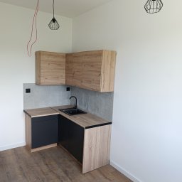Usługi remontowe malowanie, panele, płytki. - Narożna, jasna kuchnia z drewnianymi szafkami i czarnym zlewem. Widoczne podłączenie elektryczne i lampa wisząca. Nowoczesne wykończenie wnętrza.