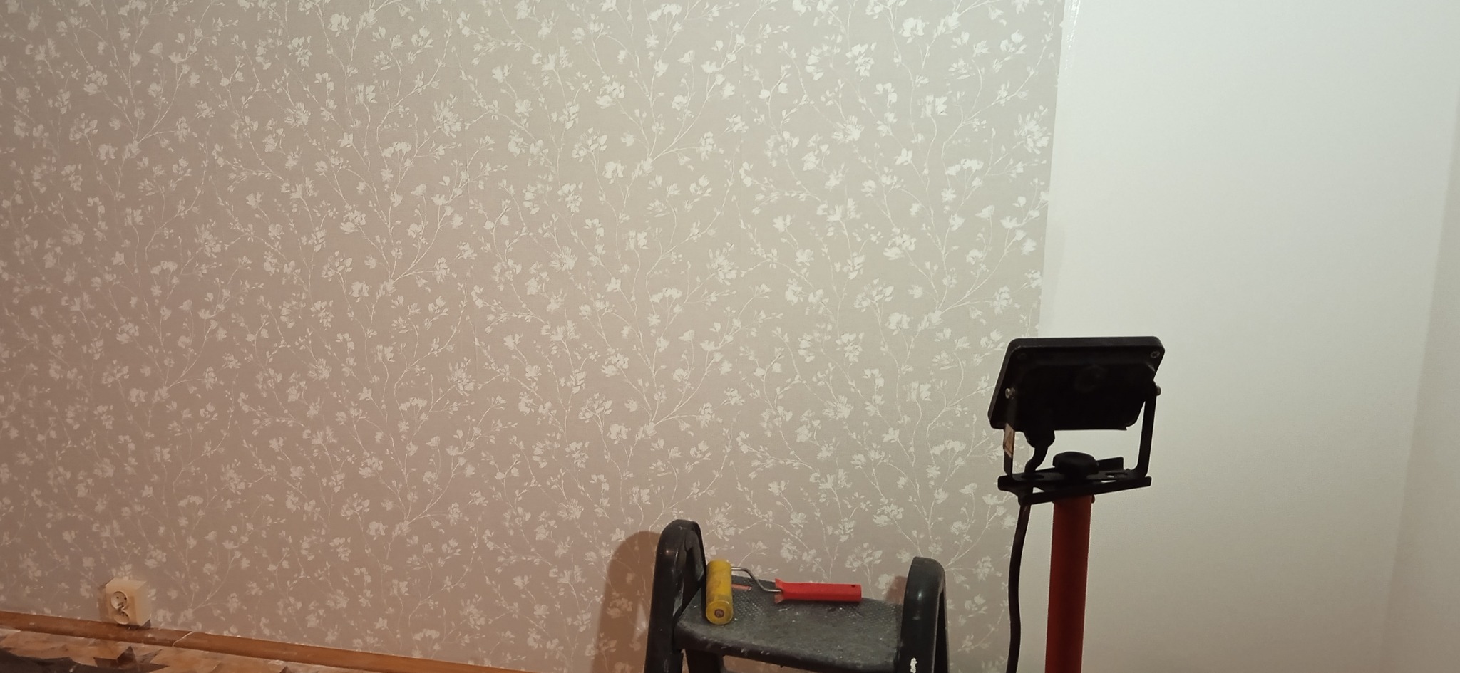 Ściana po częściowym pomalowaniu z wzorem kwiatowym, obok drabina z wałkiem i lampa robocza na statywie, widoczny fragment gniazdka elektrycznego przy podłodze.