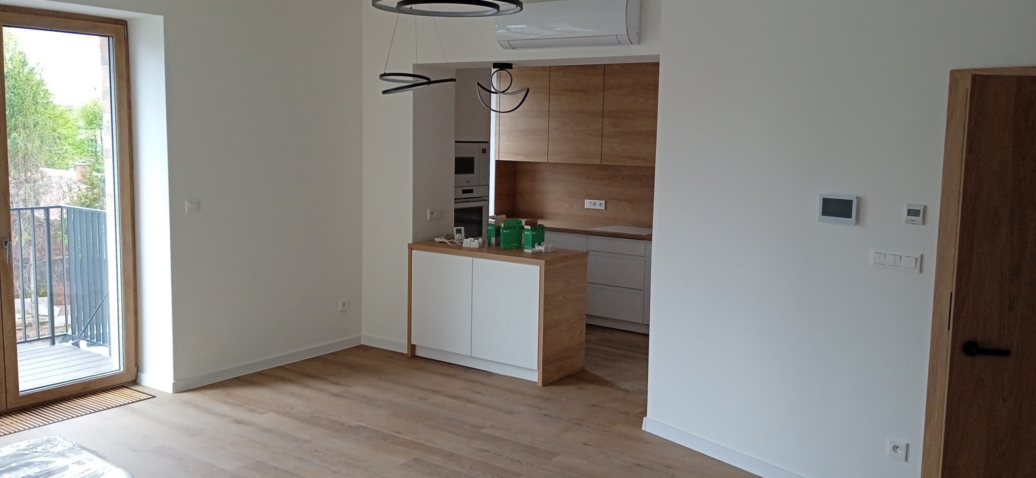 Jasne wnętrze mieszkania z podłogą z paneli w odcieniach drewna, widok na balkon i otwartą kuchnię z drewnianymi szafkami, nowoczesne oświetlenie.