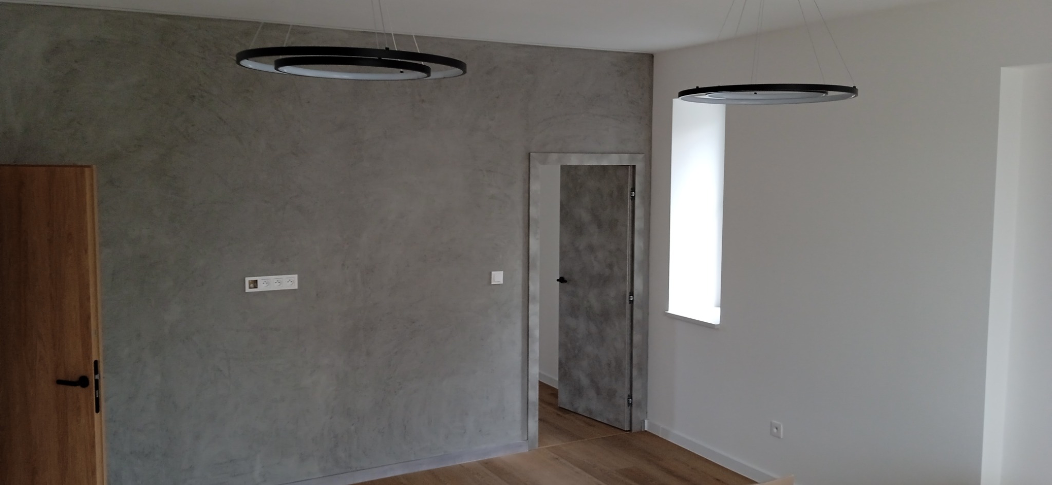 Ściana w szarym kolorze z efektem dekoracyjnym, drewniana podłoga, nowoczesne lampy wiszące w kształcie okręgów, widoczne gniazdka elektryczne i drewniane drzwi.