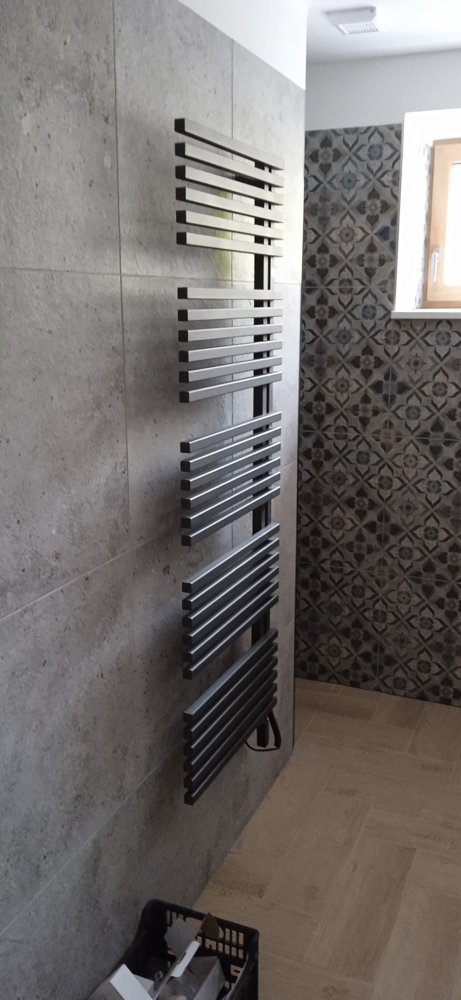 Nowoczesny, metalowy grzejnik drabinkowy w łazience z szarymi płytkami ściennymi i dekoracyjnymi płytkami w strefie prysznicowej, widoczne akcesoria montażowe w koszu na podłodze.