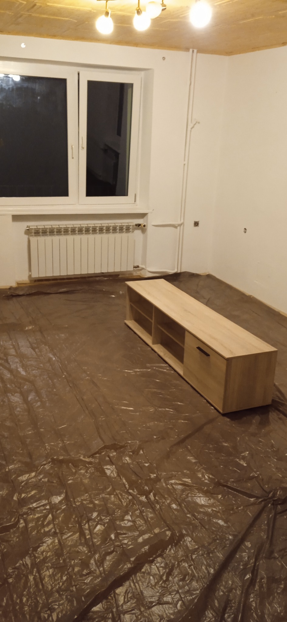 Pokój w trakcie przygotowań do prac wykończeniowych, podłoga zabezpieczona folią ochronną, widoczny grzejnik i częściowo umeblowany szafką RTV z jasnego drewna.
