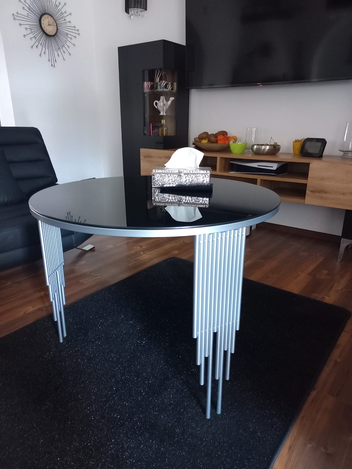 Czarny stół z okrągłym szklanym blatem i metalowymi nogami z rurek, stojący na czarnym dywanie w nowoczesnym salonie.