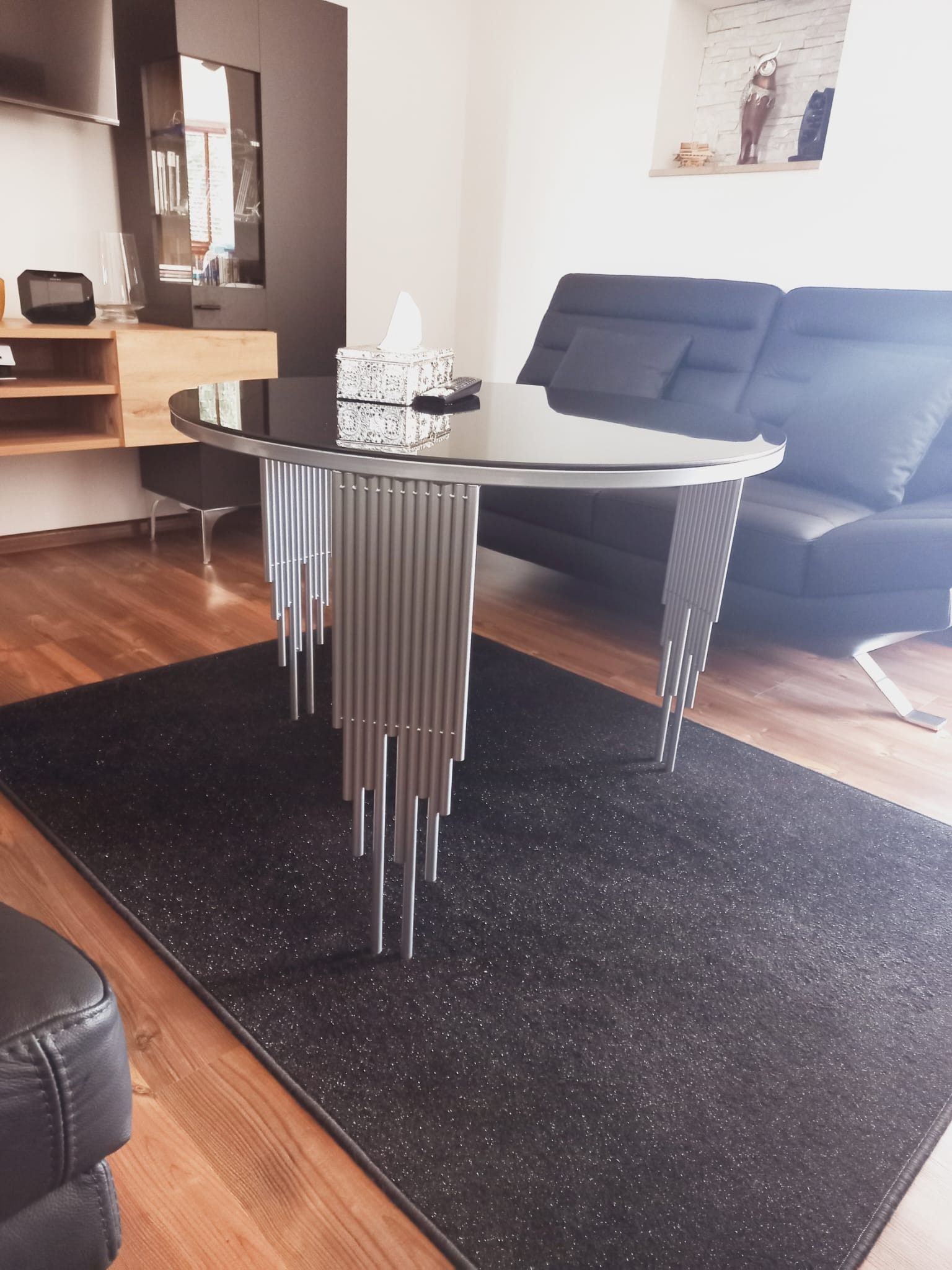 Okrągły stolik kawowy z czarnym szklanym blatem i srebrnymi, rurkowymi nogami stoi na ciemnoszarym dywanie w salonie z drewnianą podłogą i szarą sofą.