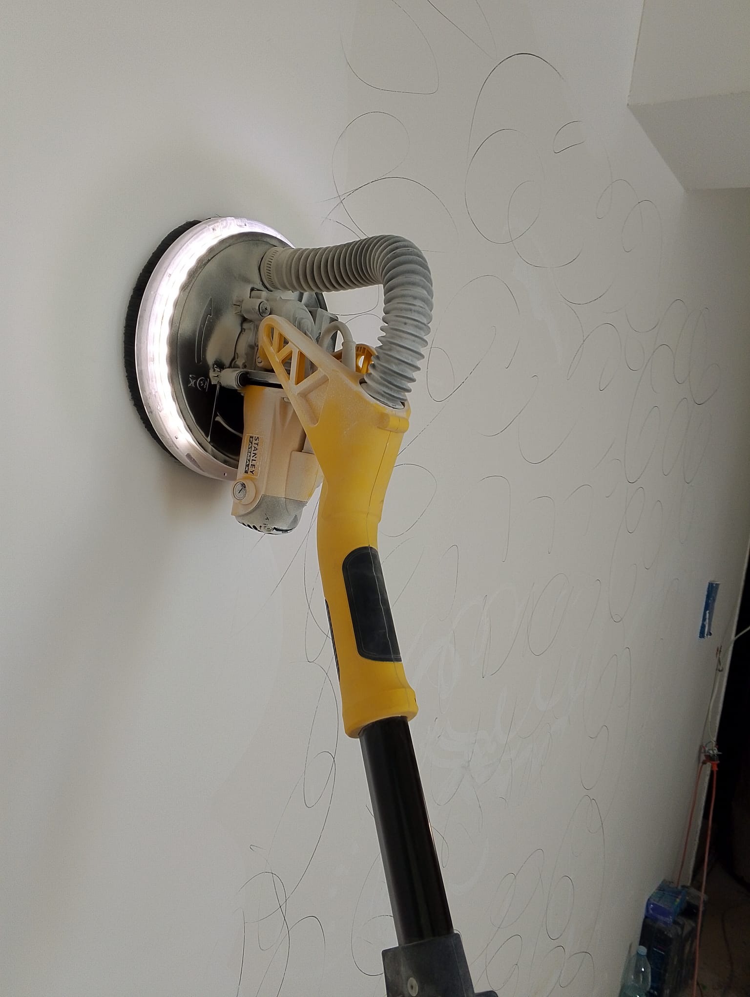 Szlifierka do gładzi gipsowych Stanley Fatmax z oświetleniem LED na ścianie z widocznymi śladami szlifowania. W tle kabel i butelka.