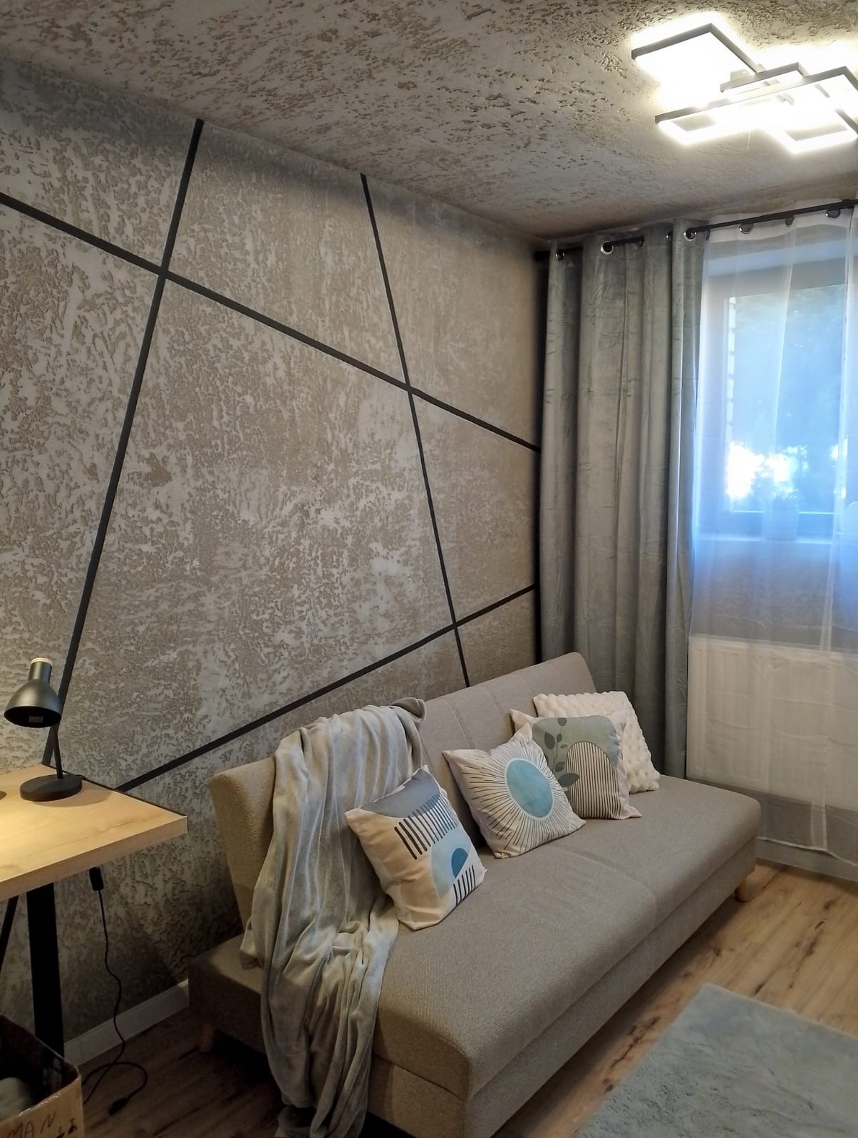 Stylowy pokój z teksturowaną ścianą zdobioną czarnymi liniami, szarą sofą z poduszkami i drewnianym biurkiem z lampką. Okno z zasłonami w tle.