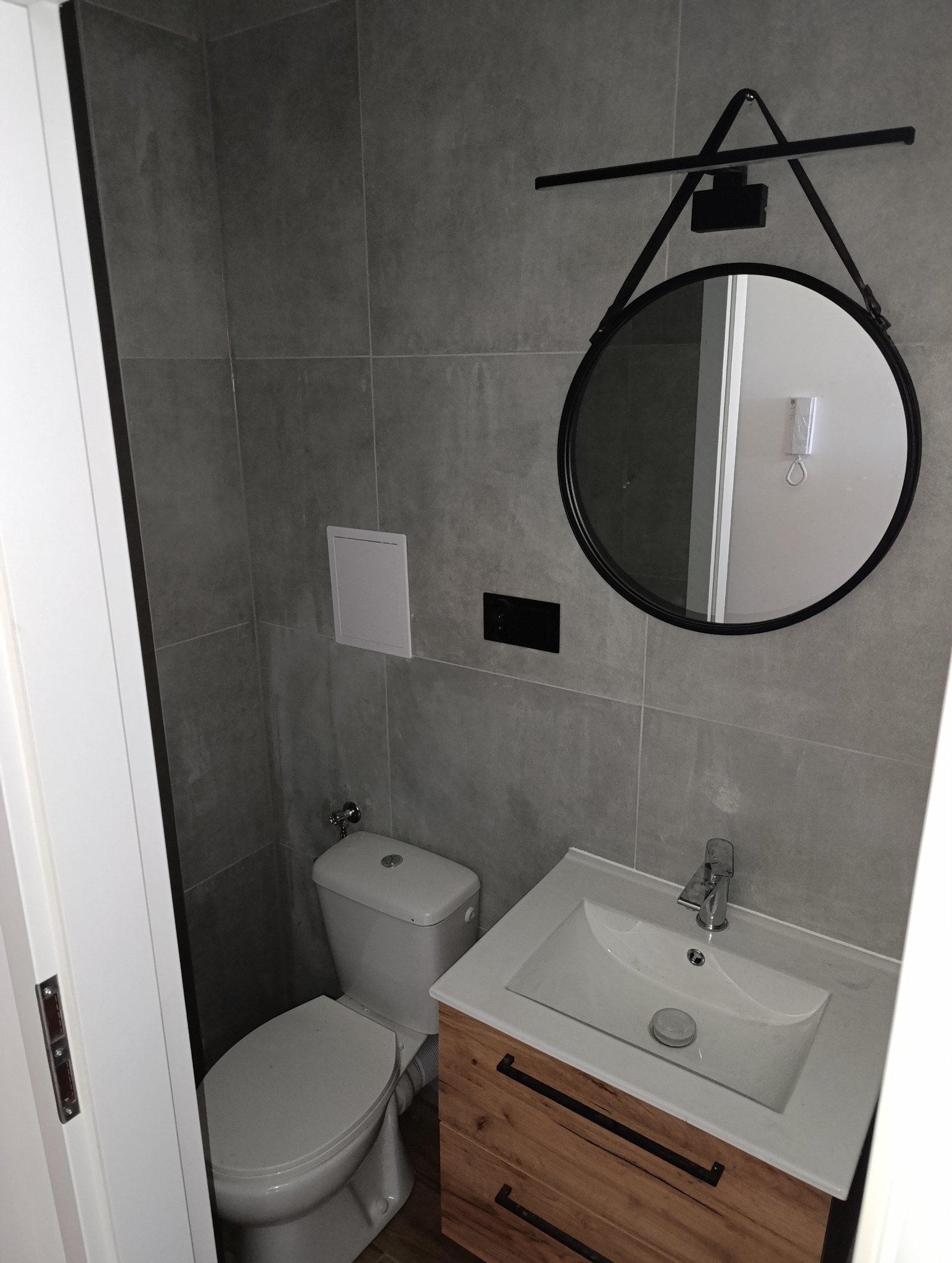 Szara łazienka z okrągłym lustrem z oświetleniem, toaletą i umywalką z szafką z drewnianymi szufladami i czarnymi uchwytami. Minimalistyczny design.