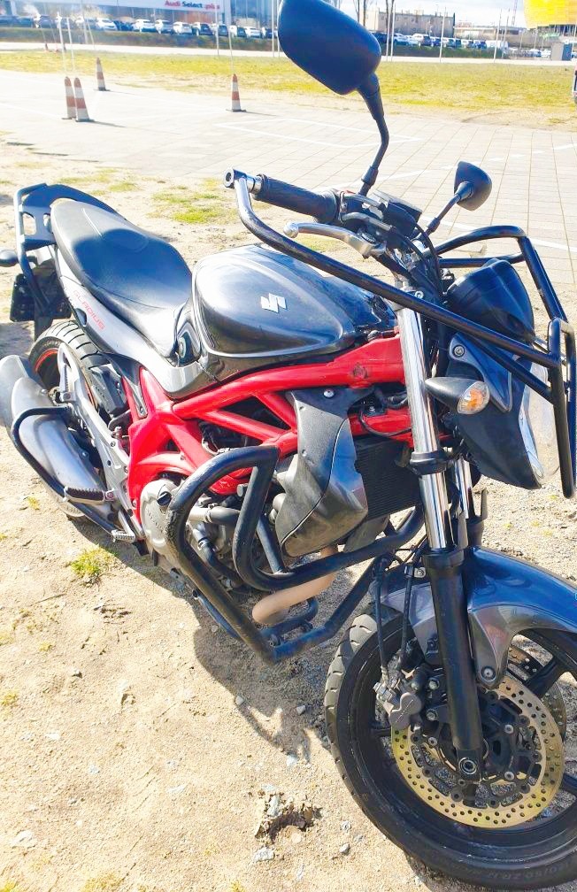 Motocykl Suzuki Gladius z czerwonym stelażem ochronnym, stojący na placu manewrowym z pomarańczowymi pachołkami w tle.