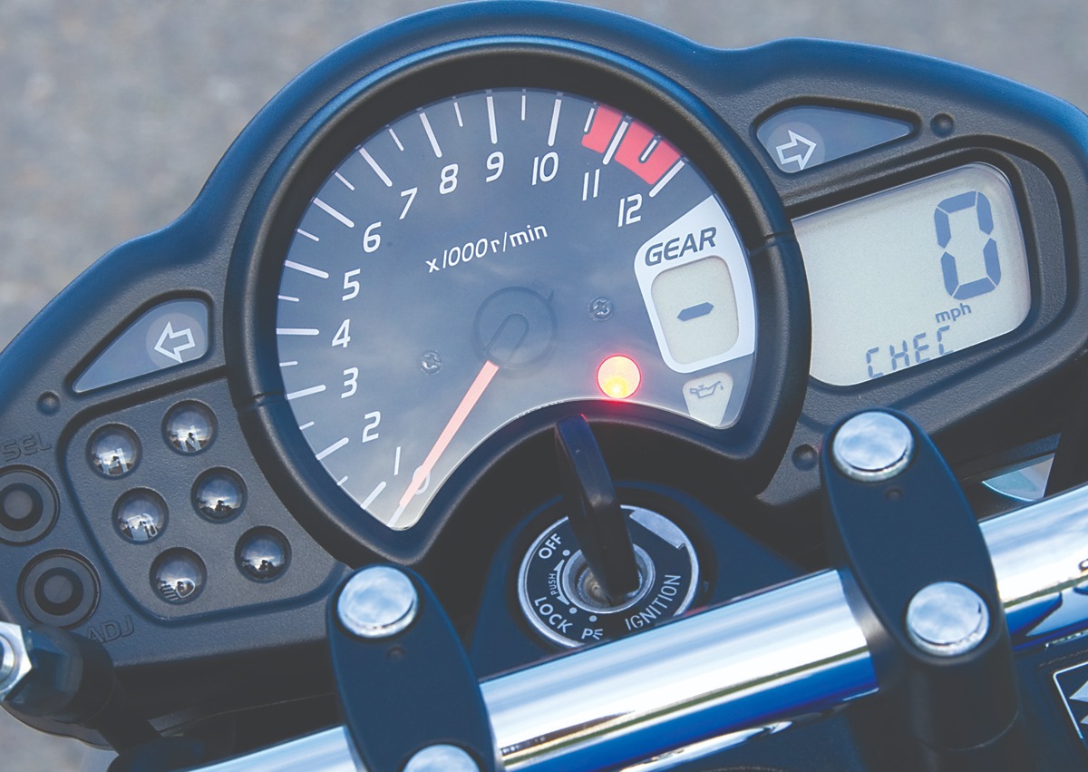 Zbliżenie na deskę rozdzielczą motocykla z włączonym zapłonem, wskazówka obrotomierza na 5000 obr./min, wyświetlacz pokazuje 0 mph, kontrolka oleju się świeci.