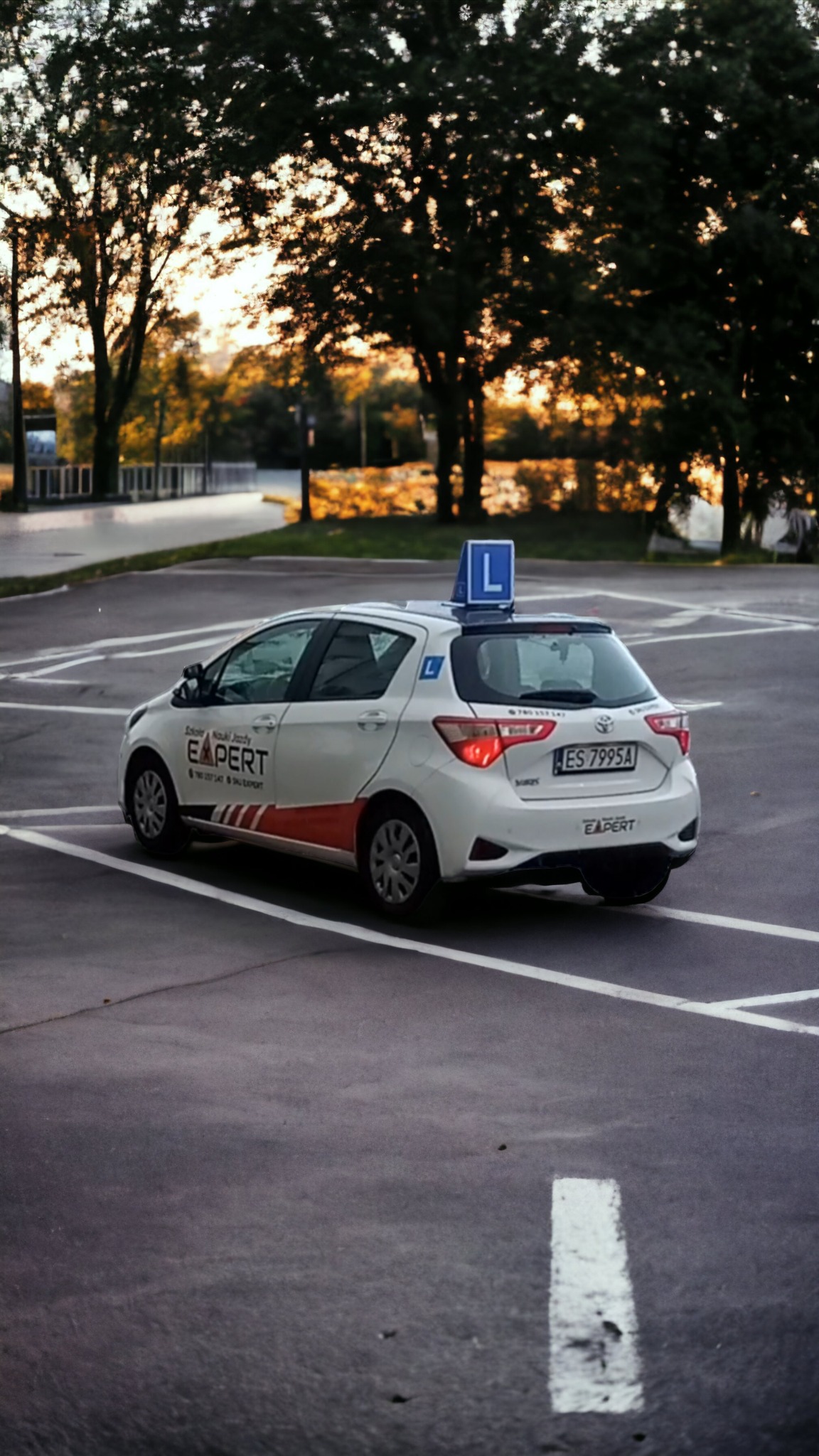 Biały samochód z literą 'L' na dachu, zaparkowany na parkingu z widocznym logo firmy EXPERT po bokach, w tle drzewa i zachodzące słońce.