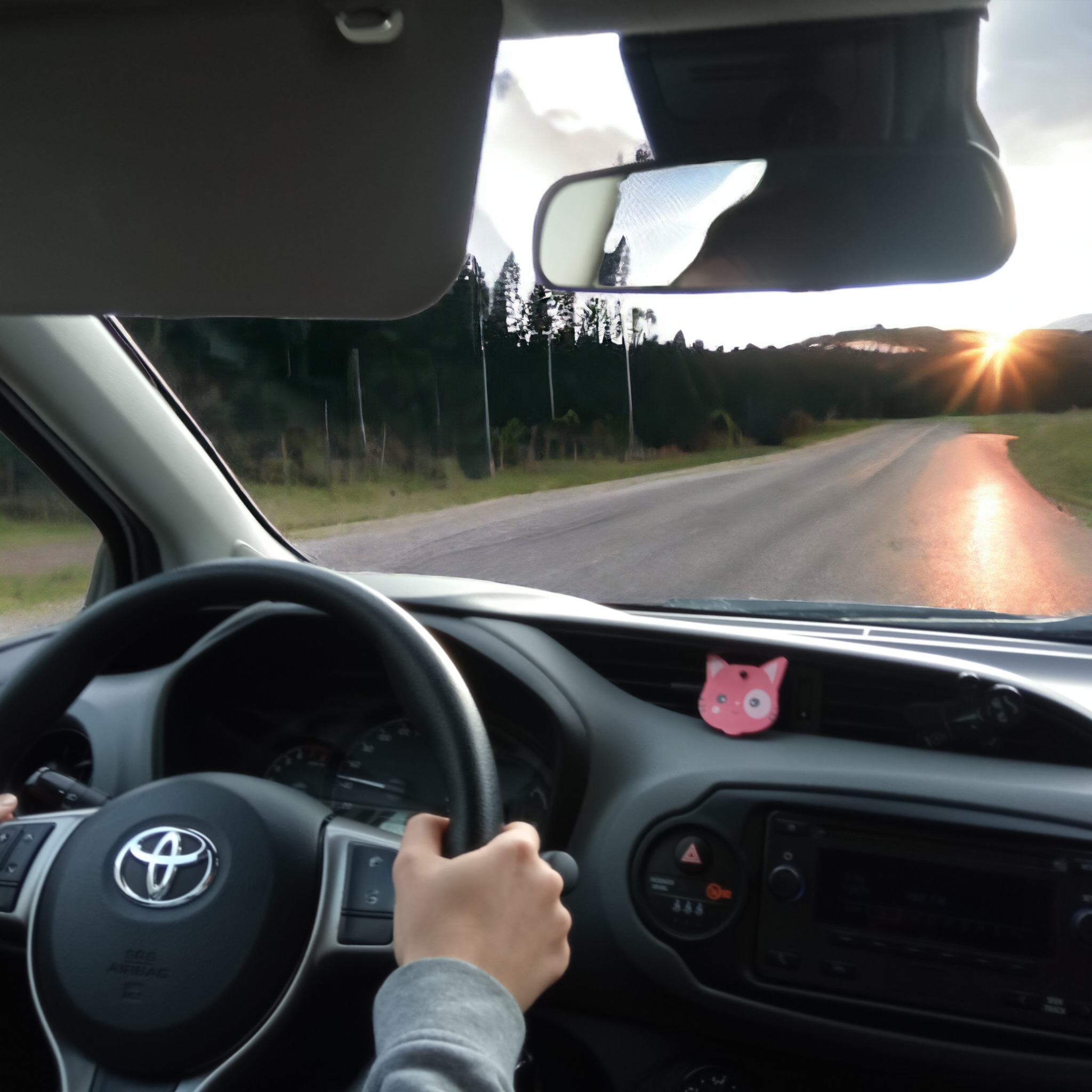Wnętrze samochodu Toyota Yaris z widokiem na drogę i zachodzące słońce, ręka na kierownicy, mała różowa zawieszka w kształcie kota na desce rozdzielczej.