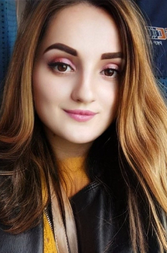 Portret kobiety z brązowymi oczami i włosami w odcieniach brązu i blondu, z delikatnym makijażem w odcieniach różu, ubrana w czarną skórzaną kurtkę z żółtą bluzką pod spodem, na rozmytym tle.