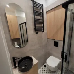 Nowoczesna łazienka z owalnym lustrem, czarną umywalką nablatową, grzejnikiem drabinkowym, szafkami wiszącymi z jasnego drewna i toaletą podwieszaną na tle szarej płytek ściennych i podłogi...