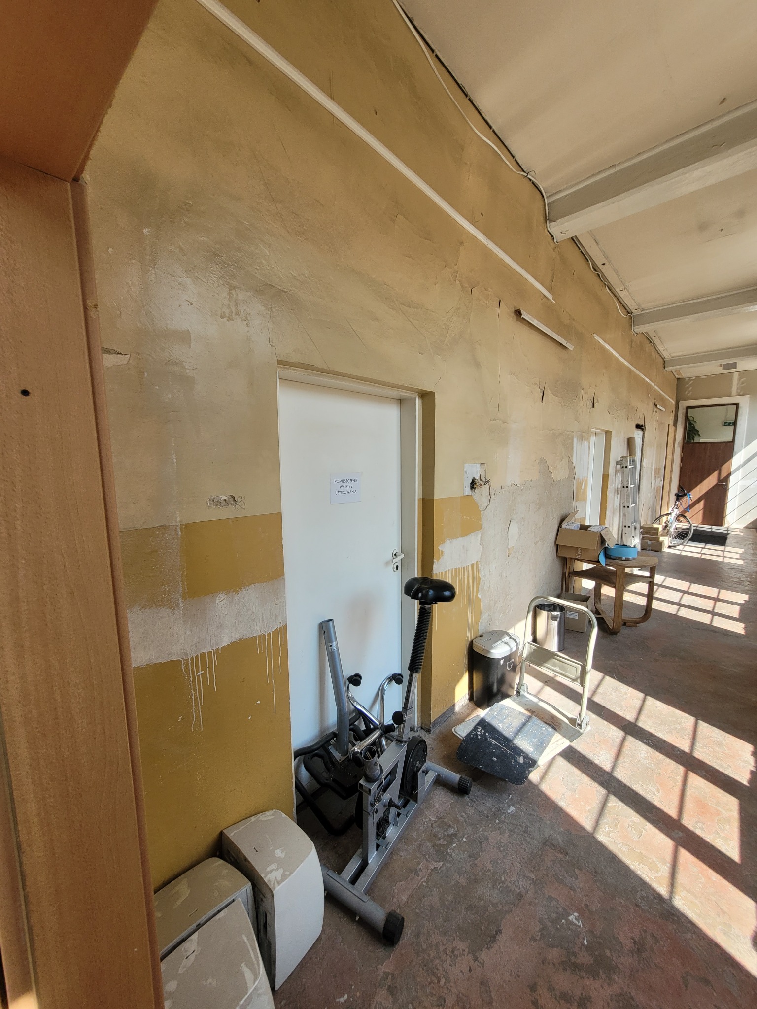 Korytarz biurowy w trakcie odświeżania: widoczne ślady po malowaniu, przygotowane ściany, sprzęt do ćwiczeń i drabina w tle.