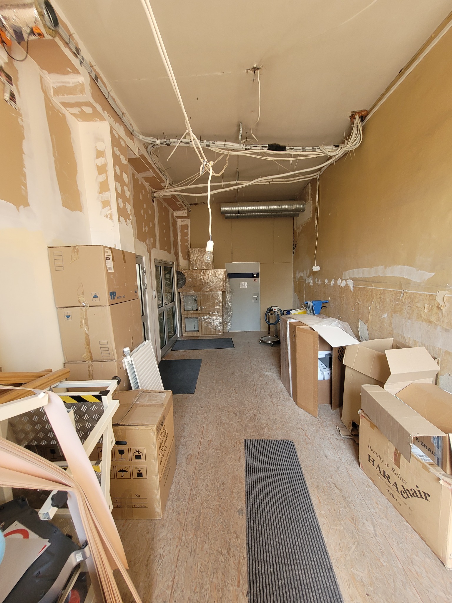 Wnętrze lokalu w trakcie prac remontowych: surowe ściany z widocznymi śladami szpachlowania, niezabezpieczone przewody elektryczne pod sufitem, kartony z wyposażeniem i narzędziami na podłodze.