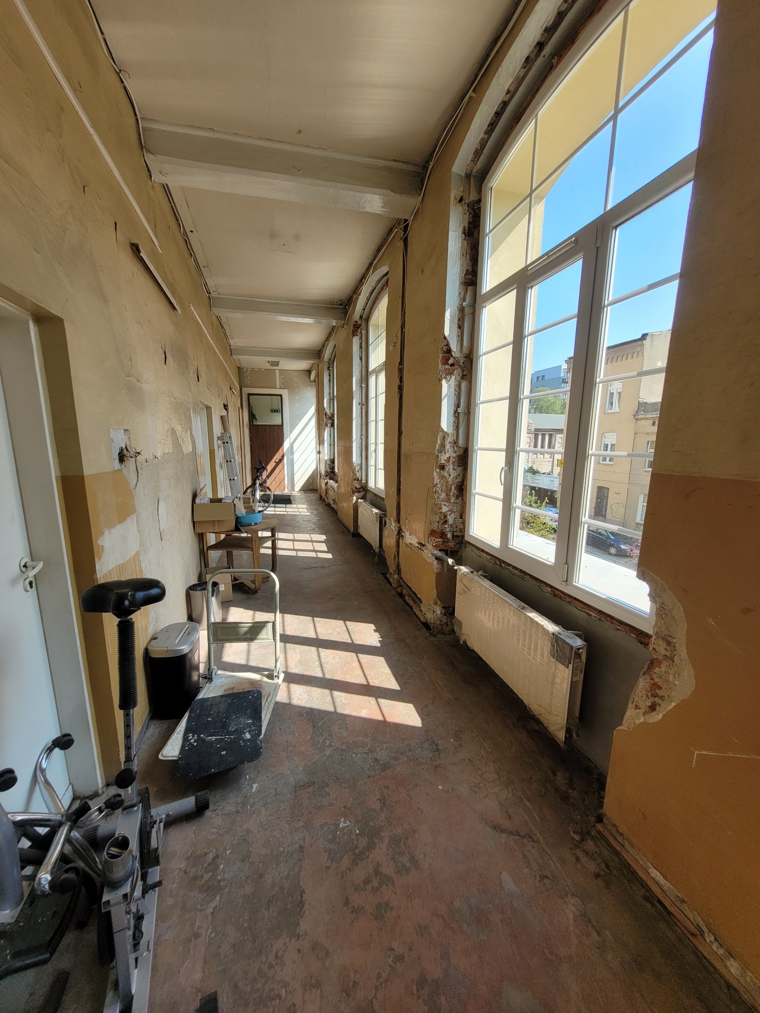 Korytarz biurowy w trakcie remontu: przygotowane ściany do malowania, widoczne fragmenty cegieł, nowe okna z widokiem na budynki, stare grzejniki zabezpieczone folią, sprzęt remontowy w rogu.