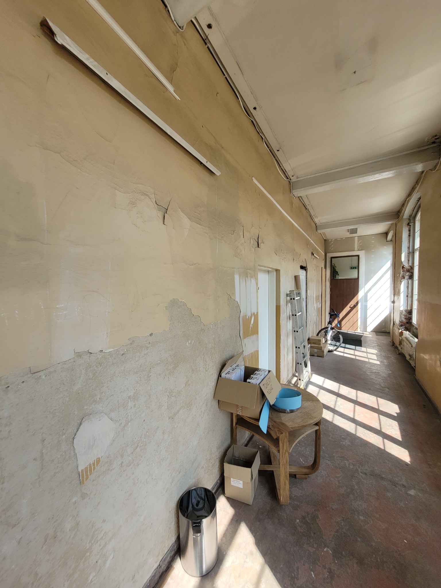 Wnętrze korytarza w trakcie remontu z widocznymi odpadającymi fragmentami starej farby na ścianach, metalowym koszem na śmieci, stolikiem z kartonami i drabiną opartą o ścianę w głębi.