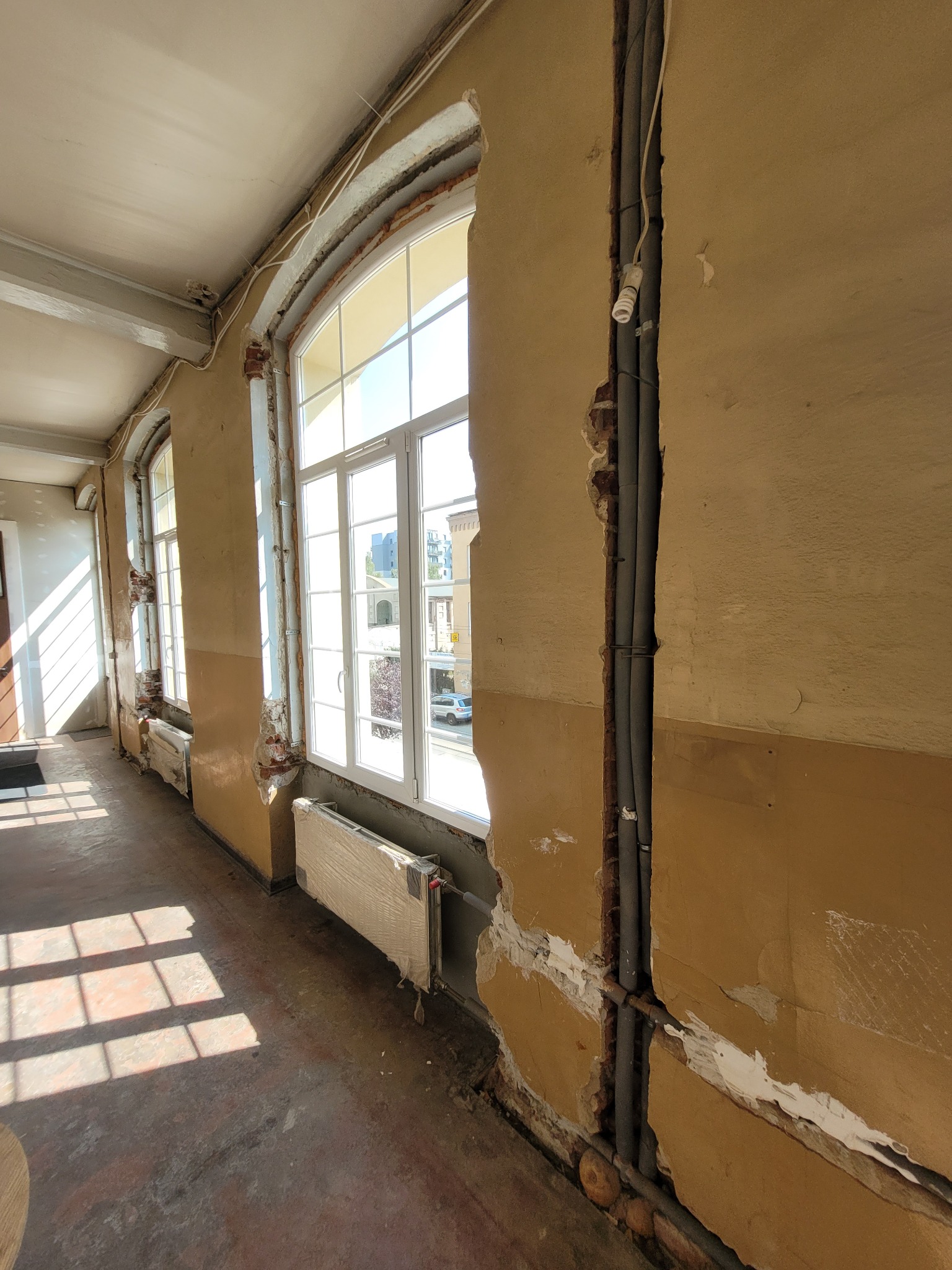 Wnętrze w trakcie remontu: odsłonięte fragmenty cegieł wokół okien z białymi ramami, widoczne instalacje w wyżłobieniach ściany, grzejnik zabezpieczony folią, ślady po starych powłokach na ścianach...